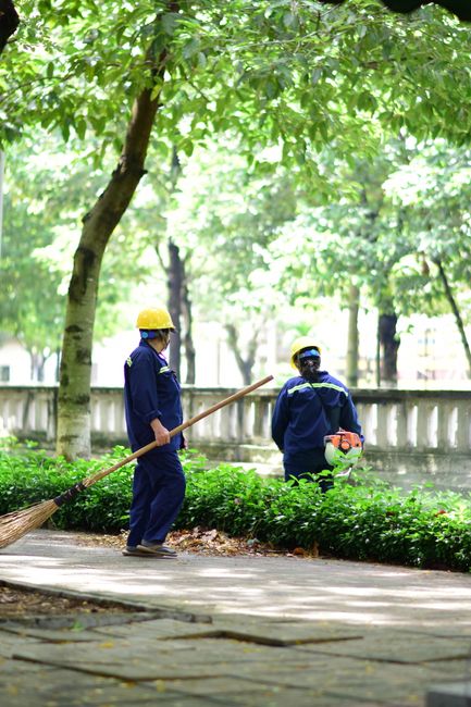 Two outdoor workers wearing blue uniforms and yellow safety helmets are engaged in garden maintenance along a paved pathway. One worker is holding a broom and appears to be sweeping fallen leaves or d