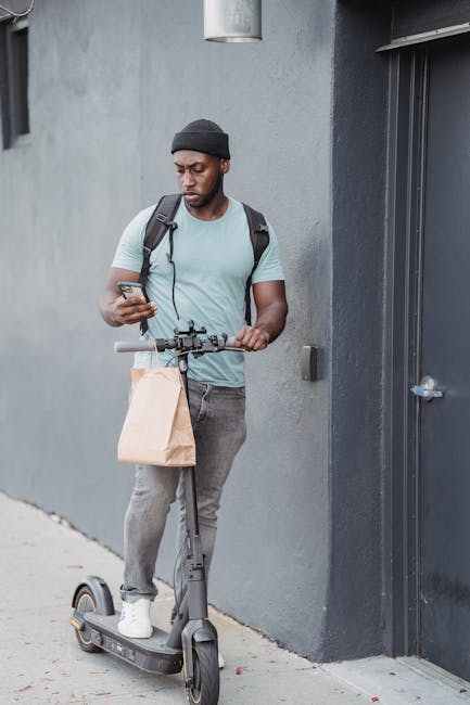 A young man with dark skin, wearing a black beanie, light blue t-shirt, and grey jeans, stands next to a grey wall and a black door while looking at his smartphone. He carries a black backpack over hi